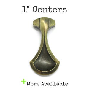 May include: A brass-toned metal cabinet pull with a flared, bell-like shape and a dark, recessed center. The text 1" Centers is printed above the object, and the text + More Available is printed below the object on a plain white background.