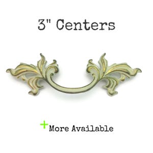 May include: A decorative drawer pull with a cream-colored finish and gold accents. The pull has a curved handle and ornate, leaf-like details. The text "3" Centers" is above the pull, and "More Available" is below.