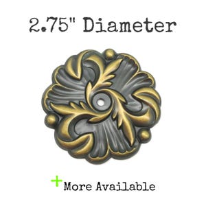 May include: A round, metallic decorative medallion with a dark grey base and raised, polished brass-toned leaf patterns. It features a central hole and a 2.75" diameter. The text "2.75" Diameter" and "+ More Available" appear in the image.