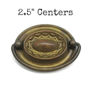 May include: An oval brass drawer pull with a rope-style border and a central raised oval medallion featuring decorative scalloped edges. The hardware includes a swinging bail handle and is labelled with the text 6.35 cm Centers above the object.