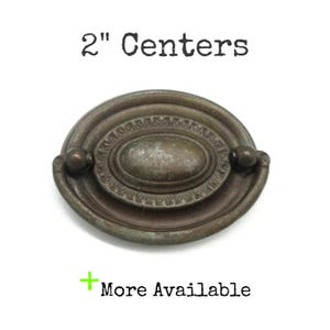 May include: An oval-shaped metal drawer pull with a dark, weathered bronze finish. The hardware features a central oval plate with a decorative border and a hinged drop handle. The text 2" Centers and + More Available is visible.