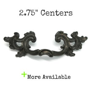 May include: A dark bronze-toned metal drawer pull with ornate, swirling scrollwork details. The text 2.75" Centers is printed above the handle, and a green plus sign with the text More Available is printed below the handle.