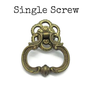May include: A brass-toned metal drawer pull featuring an ornate, decorative backplate with a floral motif and a swinging drop handle. The text "Single Screw" is printed in black above the hardware, indicating the mounting style for this piece.