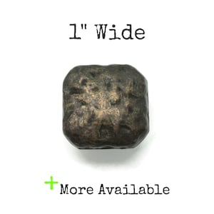 May include: A square-shaped metal button with a dark, weathered bronze finish and a textured, uneven surface. The text 1" Wide is printed above the button, and the text + More Available is printed below the button.