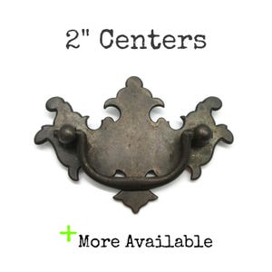May include: A dark metal drawer pull with a decorative backplate and a swinging bail handle. The text 2" Centers is printed above the hardware, and the text + More Available is printed below the hardware on a plain white background.