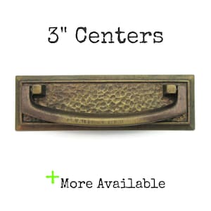 May include: A rectangular, antique brass drawer pull with a hammered texture and a curved handle. The text "3" Centers" is above the pull, and "More Available" is below. The drawer pull measures 3 inches.