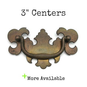 May include: Antique brass drawer pull with a decorative design. The pull has a curved handle and ornate details. The text "3" Centers" is above the pull, and "More Available" is below. The pull measures 3 inches between the screw holes.