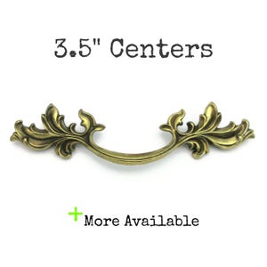 May include: Ornate brass-toned drawer pull with a curved handle and decorative leaf accents. The text "3.5" Centers" is above the pull, and "More Available" is below. The pull measures 3.5 inches (8.89 cm) between the screw holes.