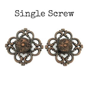 May include: Two ornate, bronze-coloured, decorative cabinet knobs with a single screw each. The knobs have a square shape with a detailed, floral design. The text "Single Screw" is visible above the knobs.