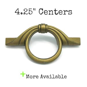 May include: A brass-toned metal drawer pull featuring a circular ring handle attached to a horizontal mounting plate. The text 4.25" Centers is printed above the hardware, and the text + More Available is printed below the hardware.
