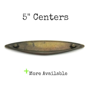 May include: An elongated, boat-shaped metal drawer pull with a dark, mottled bronze finish and pointed ends. The hardware features two small circular mounting points. Text above reads 5" Centers and text below reads + More Available.