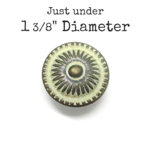 May include: A round cabinet knob with a cream-colored center and a dark bronze-toned sunburst pattern. The edge features a beaded border. Text above the knob reads: Just under 1 3/8" Diameter. The knob has a distressed finish with dark metal showing.