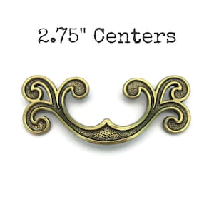 May include: Antique brass-toned drawer pull with ornate scrollwork design. The pull measures 2.75 inches between the centers of the mounting holes. The text "2.75" Centers" is printed above the pull.
