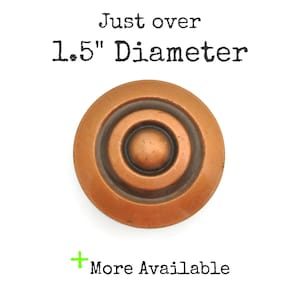 May include: A round, copper-colored knob with concentric circles and a central raised button. The text "Just over 1.5" Diameter" is above the knob, and "More Available" is below.