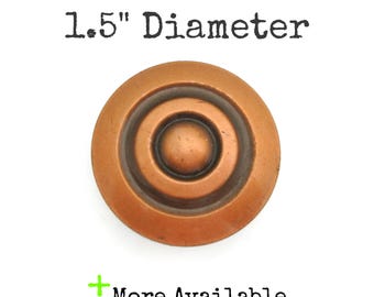 May include: A round, copper-colored knob with concentric circles and a central raised button. The text "Just over 1.5" Diameter" is above the knob, and "More Available" is below.