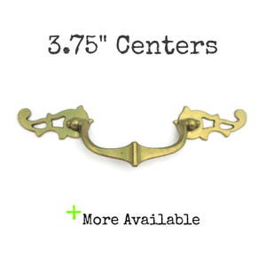 May include: A brass-toned drawer pull with a decorative design. The pull has a curved handle and measures 3.75 inches between the centers of the mounting holes. The text "3.75" Centers" is above the pull, and "More Available" is below.