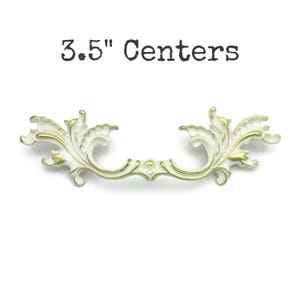 May include: White and gold decorative drawer pull with an ornate, floral design. The text "3.5" Centers" is above the pull. The pull measures 3.5 inches between the centers of the mounting holes. This hardware is suitable for cabinets and drawers.
