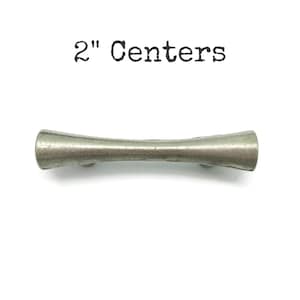 May include: A silver-toned cabinet pull with a curved, hourglass shape. The text "2" Centers" is printed above the pull. The hardware is approximately 2 inches in length.