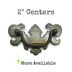 May include: A dark bronze-toned metal drawer pull with a curved handle and a decorative backplate featuring ornate cutouts. The text 2" Centers is printed above the hardware, and the text + More Available is printed below the hardware.