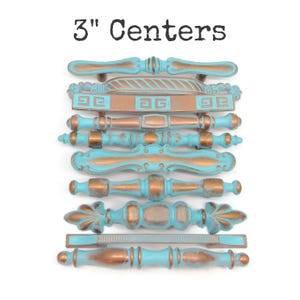 May include: A collection of nine drawer pulls with 3" centers, featuring a distressed finish with turquoise blue paint over a copper-toned metal base. Each handle displays a unique shape, ranging from straight bars to ornate, curved designs.
