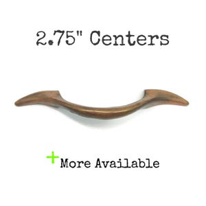 May include: A curved, bronze-toned metal drawer pull with a smooth finish. The text 6.99 cm Centers is printed above the handle, and a green plus sign with the text More Available is printed below the handle on a plain white background.