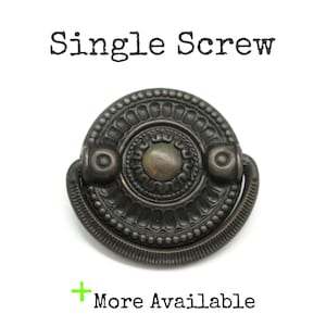 May include: A dark bronze-toned metal drawer pull with a circular backplate featuring a beaded edge and a central dome. A semi-circular drop handle is attached to the backplate. The text "Single Screw" and "+ More Available" is visible.