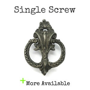 May include: A dark silver-toned metal drawer pull with an ornate, symmetrical design featuring a central vertical element and a circular ring. The text "Single Screw" appears above the object, and "+ More Available" is printed below it.