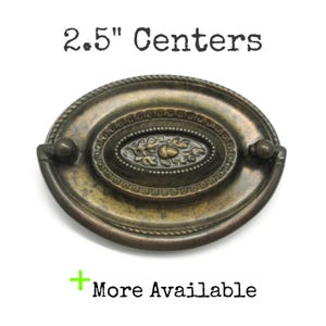 Peut inclure: Une poignée de tiroir ovale en laiton antique avec une bordure torsadée et un motif central en relief représentant un gland et des feuilles de chêne. La quincaillerie comprend une anse mobile et le texte 2.5" Centers et + More Available.