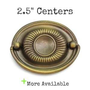 May include: An antique brass oval drawer pull with a rope-like border and a central circular design. The text "2.5" Centers" is above the pull, and "More Available" is below.