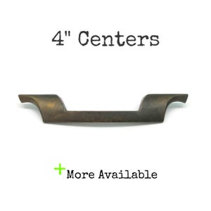 May include: A dark bronze-toned metal drawer pull with a horizontal bar and angled ends. The text 4" Centers is printed above the handle, and the text + More Available is printed below the handle in black and green.