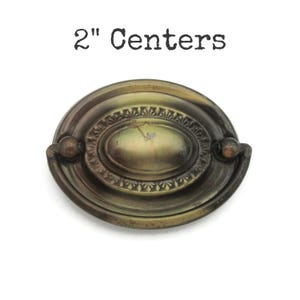 May include: Antique brass-toned drawer pull with an oval center and decorative detailing. The pull has a 2-inch center-to-center measurement and two small knobs on either side. The text "2" Centers" is above the pull.