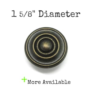 May include: A round, dark bronze-toned metal cabinet knob with a central raised dome surrounded by a textured, ridged ring. The text 1 5/8" Diameter is printed above the knob, and the text + More Available is printed below the knob.