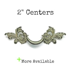 May include: A metal drawer pull with a distressed cream and dark brown finish featuring ornate scrollwork and leaf-like details. The text above reads 2" Centers and the text below includes a green plus sign followed by More Available.