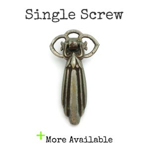 May include: A vintage-style, silver-toned drawer pull with an ornate design. The pull features a decorative top with a floral-like pattern and a teardrop-shaped handle. The text "Single Screw" is above the pull, and "More Available" is below.