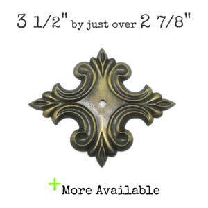 May include: A brass-toned metal decorative escutcheon plate with an ornate, symmetrical scrollwork design and a central mounting hole. The text above reads 3 1/2" by just over 2 7/8" and the text below reads + More Available.