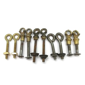 Peut inclure: Une collection de dix boulons à œil en métal avec des tiges filetées et des écrous attachés. Les pièces de quincaillerie varient en couleur, présentant des finitions en or métallique, laiton et oxydé foncé. Chaque boulon a une tête en boucle.