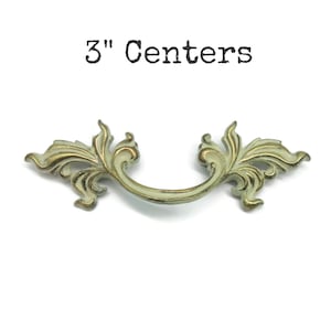 May include: An ornate, antique-style drawer pull with a cream and gold finish. The pull features a curved handle and decorative floral accents. The text "3" Centers" is printed above the pull.