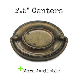 May include: An oval-shaped antique brass drawer pull with a beaded border and a drop handle. The hardware features a weathered finish and is labeled with the text 2.5" Centers and More Available, indicating a mounting distance of 2.5 inches.