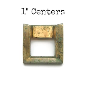 May include: A square, brass-colored metal buckle with a rectangular opening. The buckle has a weathered appearance with green and gold tones. The text "1" Centers" is printed above the buckle.