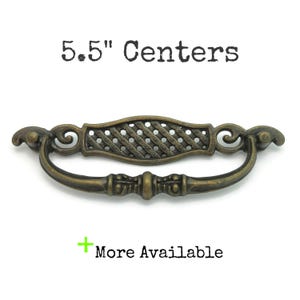 May include: Antique brass-finished drawer pull with a decorative lattice center and curved handle. The text "5.5" Centers" is above the pull, and "More Available" is below. The pull measures 5.5 inches (14 cm) between the mounting holes.
