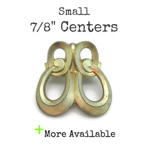 May include: A small, antique-style brass drawer pull with a decorative, symmetrical design. The text "Small 7/8" Centers" is printed above the pull. The text "More Available" is printed below the pull.