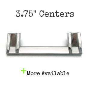 May include: A shiny, silver-toned cabinet pull with a rectangular bar and two upright supports. The text "3.75" Centers" is above the pull, and "More Available" is below. The hardware is designed for furniture.