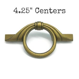 May include: A brass-toned metal drawer pull featuring a circular ring handle attached to a horizontal bar base. The text 4.25" Centers is printed above the hardware, indicating the distance between the mounting points for this furniture handle.