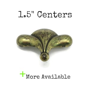 May include: A brass-toned metal drawer pull with a weathered, aged patina and a curved, symmetrical design. The hardware features a central rounded point and two side wings. The text 1.5" Centers and + More Available is visible.