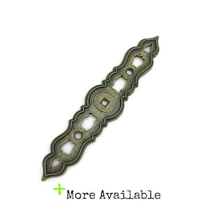 May include: An antique bronze-colored decorative drawer pull with an ornate design. The pull features a central square detail, openwork circles, and decorative edges. The text "More Available" is in the lower left corner.
