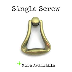 May include: A single antique brass-colored drawer pull with a teardrop shape. The pull has a rounded top and a curved handle. The text "Single Screw" is above the pull, and "More Available" is below it.
