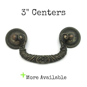 May include: Antique bronze drawer pull with a decorative floral design. The pull has a 3-inch center-to-center measurement. The text "3" Centers" and "+ More Available" are visible.