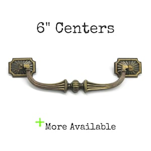 May include: An antique brass-toned metal drawer pull with a curved handle and two rectangular mounting plates featuring sunburst patterns. The text 6" Centers and + More Available is printed in black above and below the hardware piece.