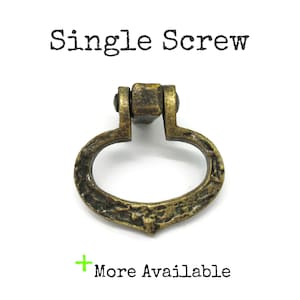May include: An antique brass-toned metal drop pull handle featuring a textured, oval-shaped ring attached to a central mounting block. The text "Single Screw" appears above the hardware, and the text "More Available" is positioned below.