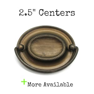May include: An oval-shaped antique brass drawer pull with a central horizontal lined pattern and a beaded border. The hardware features a swinging bail handle and is labelled with the text 2.5" Centers and More Available.
